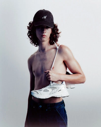 Studio shot of a shirtless adult male wearing a baseball cap and jeans, holding a white sneaker against a plain background.