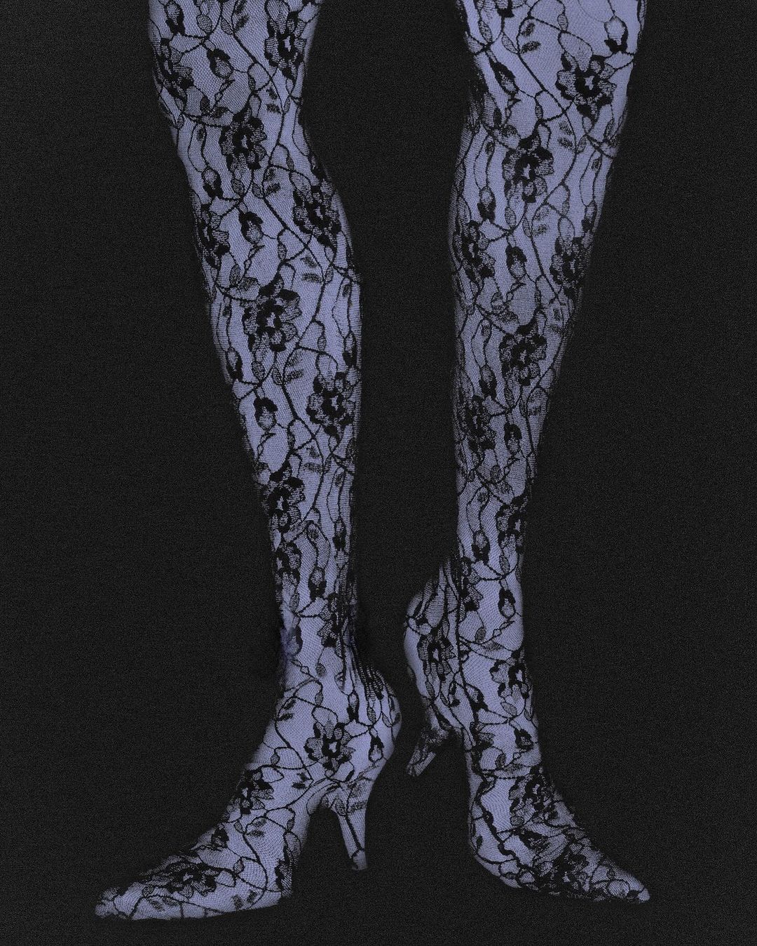 Close-up on a person's legs and feet clad in patterned lace stockings and matching heeled shoes against a dark grey backdrop.