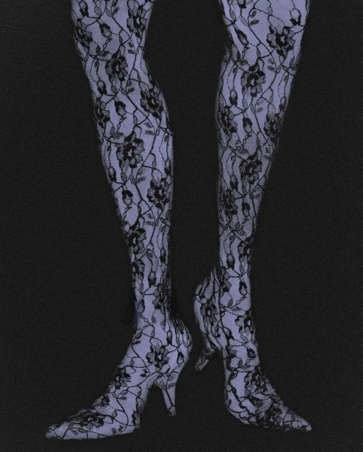 Close-up on a person's legs and feet clad in patterned lace stockings and matching heeled shoes against a dark grey backdrop.