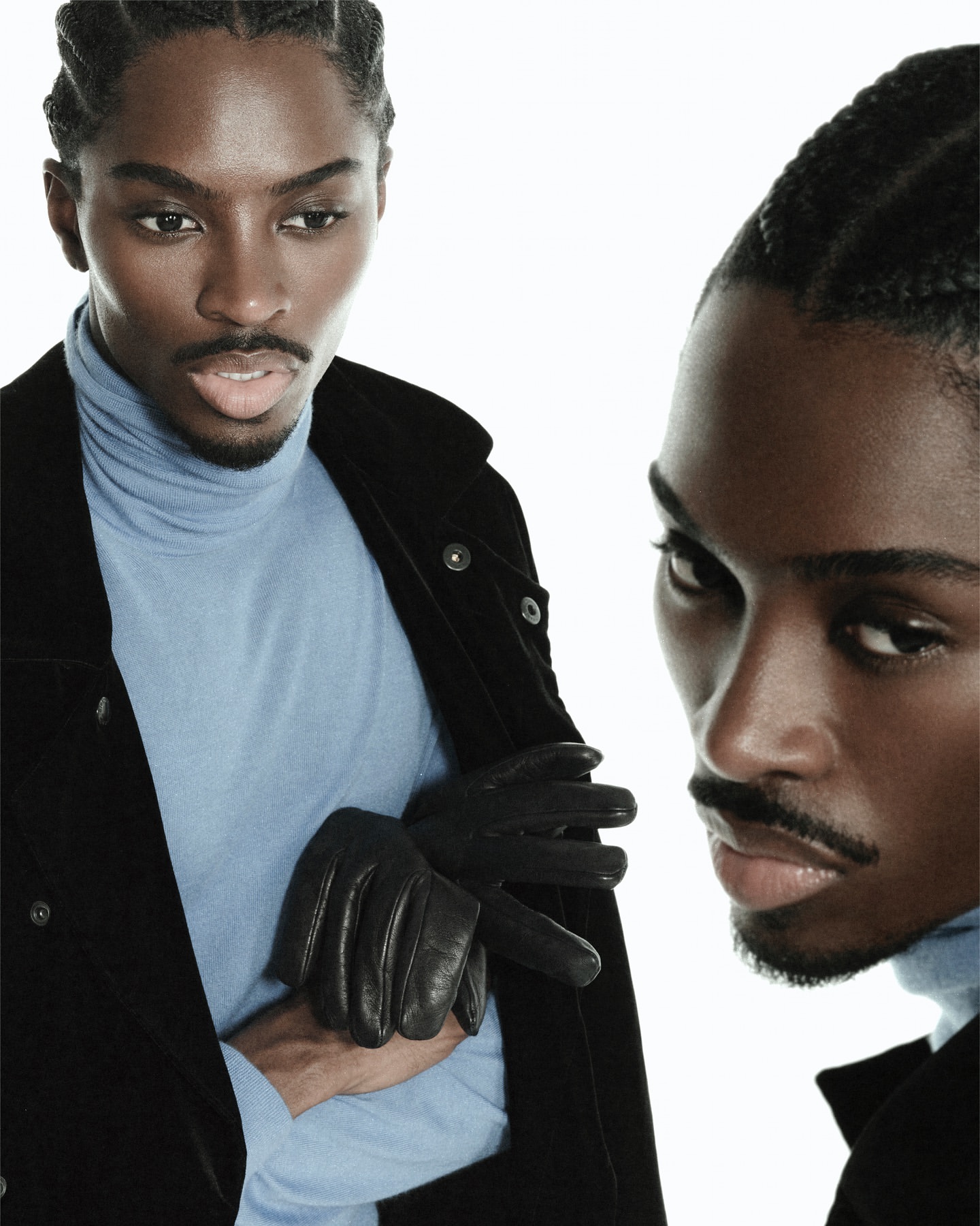 Fashion-forward portrait of a young black man with braided hair, wearing a black jacket and light blue turtleneck against a white backdrop.
