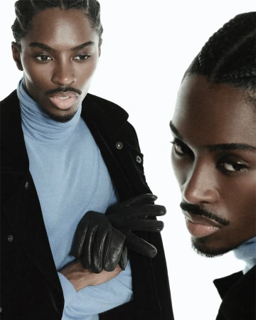 Fashion-forward portrait of a young black man with braided hair, wearing a black jacket and light blue turtleneck against a white backdrop.