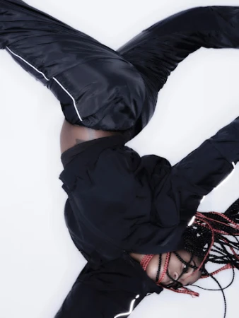 An athletic young woman in a black tracksuit does a handstand against a white backdrop, showcasing fitness and strength with style.