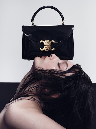 A woman with closed eyes and dark hair faces up toward a glossy black Celine handbag with a gold clasp, set against a studio backdrop.