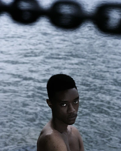 Portrait of a shirtless Black man with short hair and an intense gaze, set against a wavy water background with dark chains in the foreground.