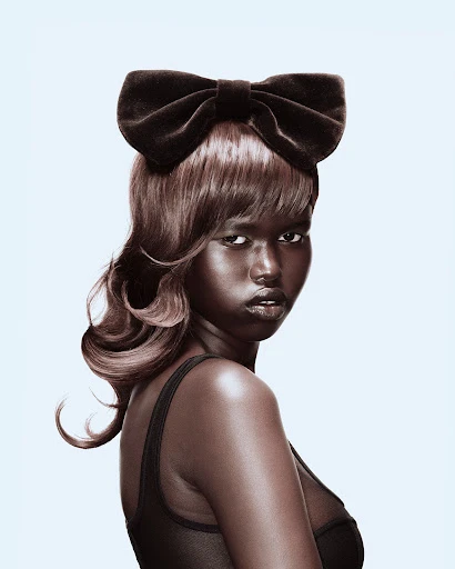 Glamorous close-up shot of a dark-skinned woman with wavy brown hair and a large black bow, wearing a black top against a light blue background.