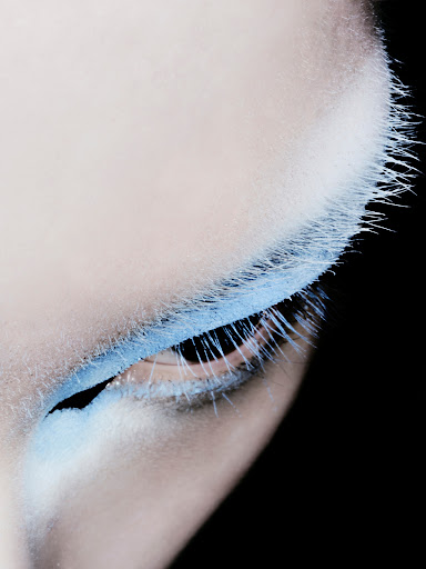 Close-up shot of an adult woman's eye with blue eyeshadow and blue-tinted eyelashes against a stark black background.