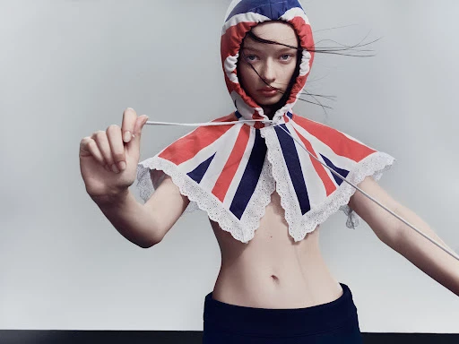Female model wearing a Union Jack themed hooded cape, posing in a photo studio against a grey backdrop with a black floor.
