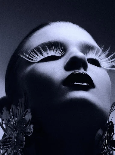 Portrait of a woman with dramatic makeup including long white eyelashes and dark lipstick, wearing large flower earrings, low angle.
