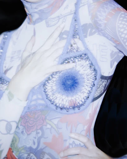 An androgynous person with a patterned top is posing. Their hand is on their chest, close to the collarbone. The background is dark.