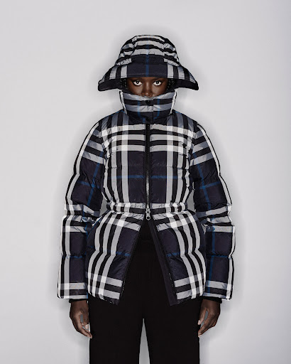 Dark-skinned woman wearing a plaid puffer jacket and matching hat against a white backdrop in a fashion editorial shot.