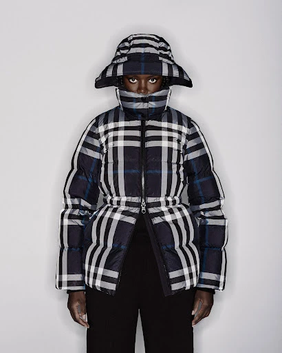 Dark-skinned woman wearing a plaid puffer jacket and matching hat against a white backdrop in a fashion editorial shot.