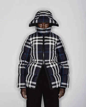 Dark-skinned woman wearing a plaid puffer jacket and matching hat against a white backdrop in a fashion editorial shot.