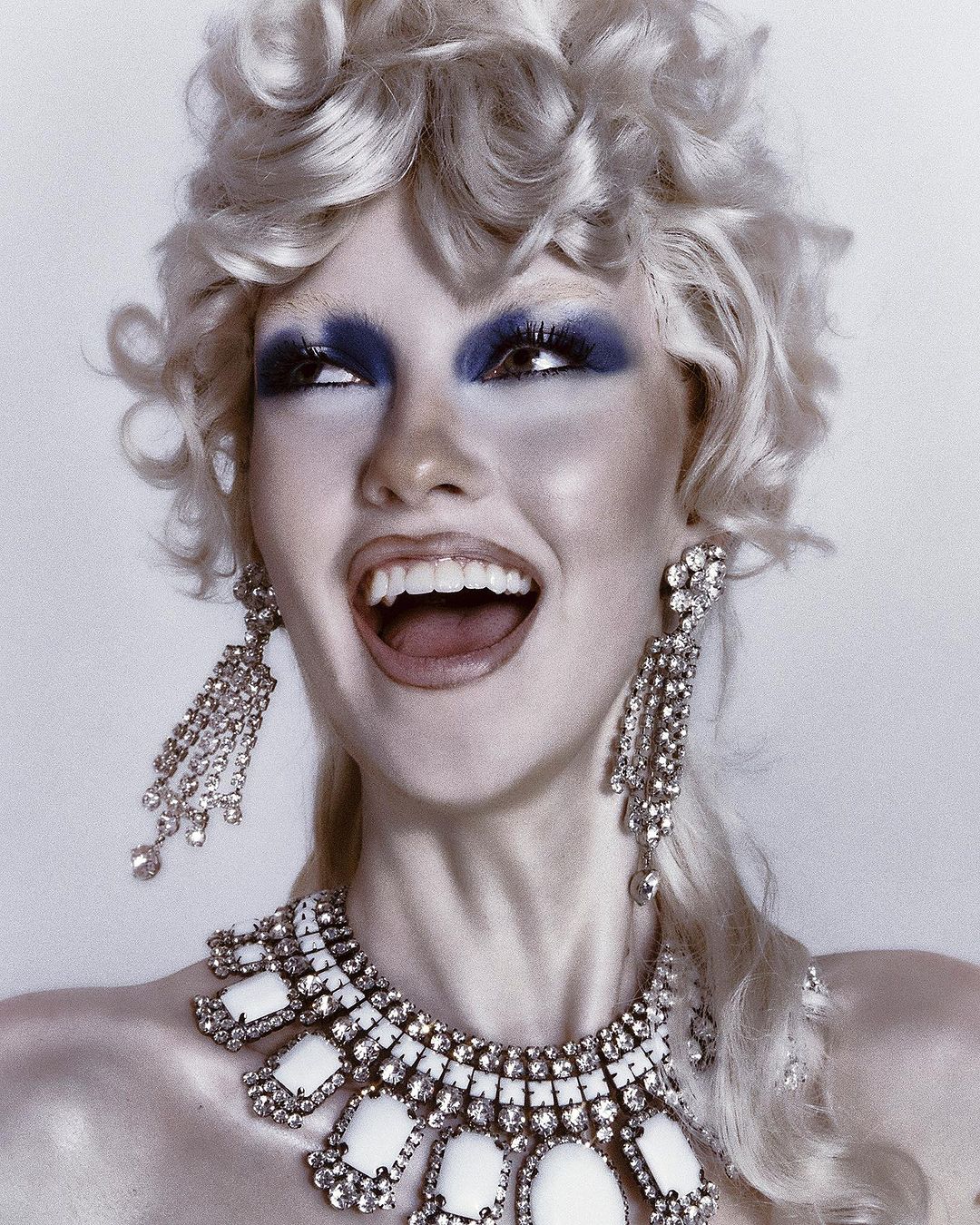 Woman with dramatic blue eye makeup, curly blonde hair, wearing statement jewelry, laughing with open mouth against a white backdrop.