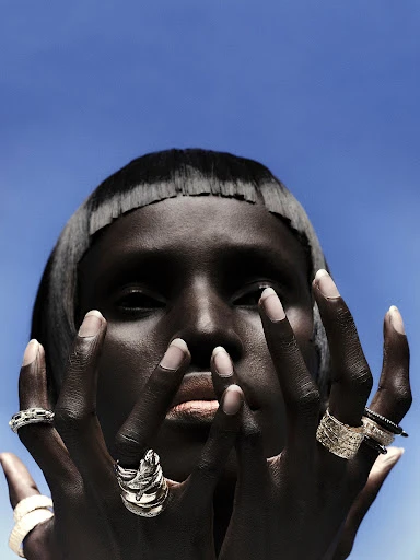 Striking portrait of a woman with dark skin and a blunt haircut, posing with heavily ringed hands against a bright, cloudless blue sky.