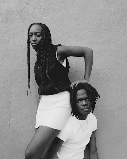 Monochrome portrait of a stylish Black woman standing over a Black man against a textured gray wall, creating a striking visual statement.