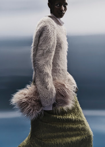An elegant, dark-skinned fashion model poses in a fur sweater and skirt, set against a soft, out-of-focus background. Vertical composition.