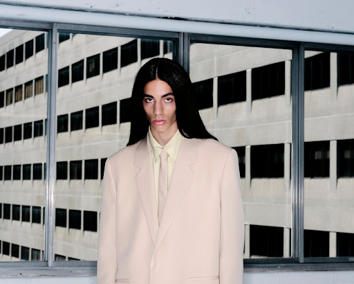 A person with long dark hair wearing a beige blazer and yellow shirt against a multi-paned window background.