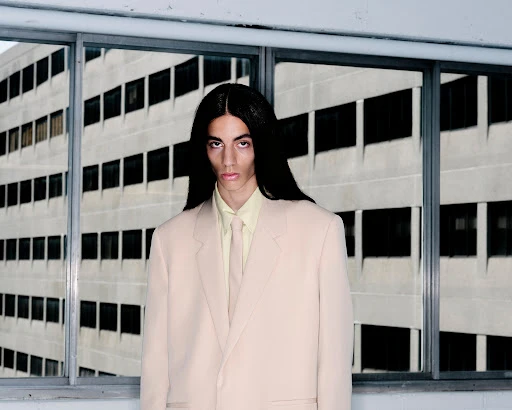 A person with long dark hair wearing a beige blazer and yellow shirt against a multi-paned window background.