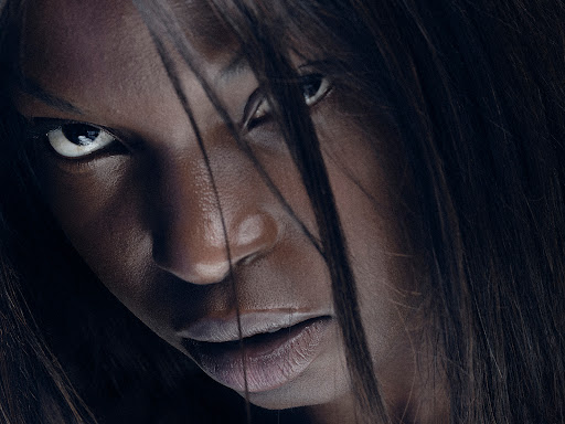 Close-up portrait of a Black adult female with dark hair partially covering her face, intense expression, against a dark background in photo studio.