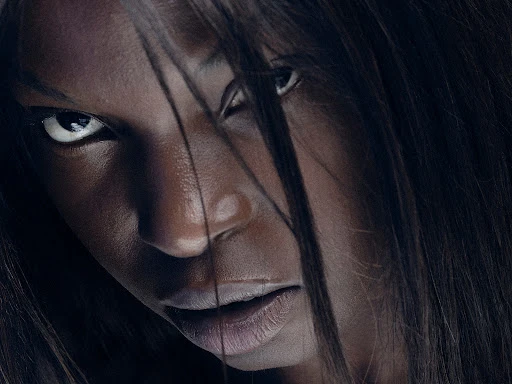 Close-up portrait of a Black adult female with dark hair partially covering her face, intense expression, against a dark background in photo studio.