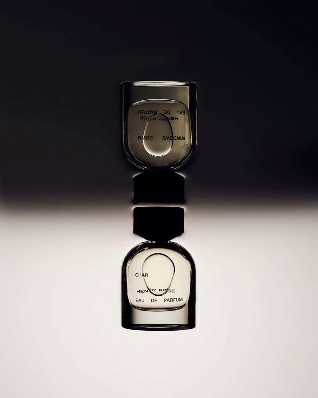Two Henry Rose Eau de Parfum bottles, stacked vertically, on a reflective surface with a dark background and soft, indirect lighting.