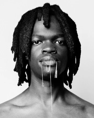 Portrait of a Black man with dreadlocks holding a delicate silver chain between his lips against a plain white background.