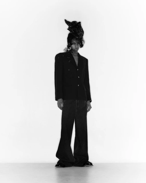 Full shot of one adult fashion model in a black blazer and dark jeans, posing in a photo studio with a black sculptural hat.