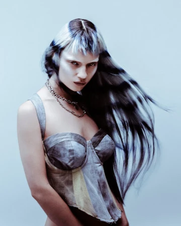 Woman with long dark and light streaked hair wearing a grey corset and chain necklace against a light background.