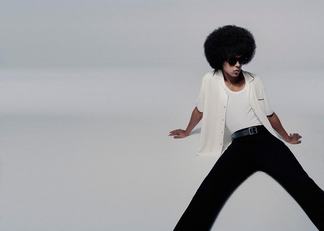 Man with afro poses in white button-down shirt and black pants against a grey backdrop. Sunglasses and black belt complete the look.