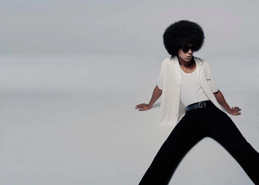 Man with afro poses in white button-down shirt and black pants against a grey backdrop. Sunglasses and black belt complete the look.