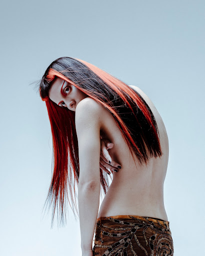 Artistic portrait of a woman with striking red and black hair, bare back, and patterned brown skirt against a light background.