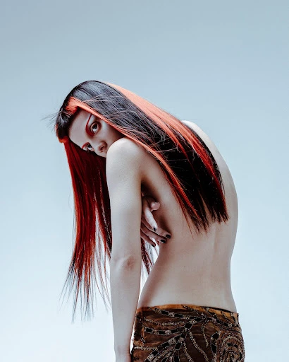 Artistic portrait of a woman with striking red and black hair, bare back, and patterned brown skirt against a light background.