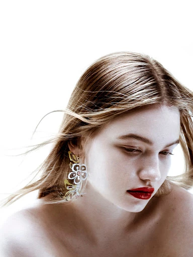 Close-up studio shot of a blonde woman with flower earrings and red lipstick in a bright white environment, showcasing fashion and beauty.