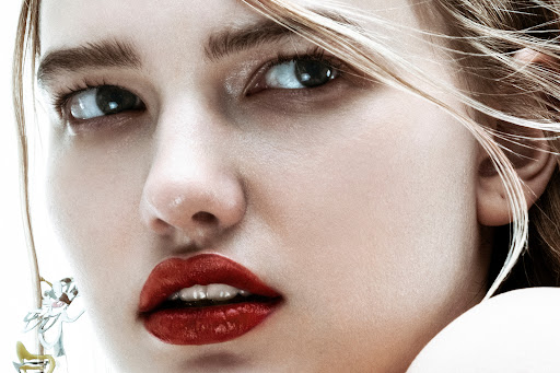 Close-up beauty portrait of a woman with glossy red lipstick and loose blonde hair against a plain light background.