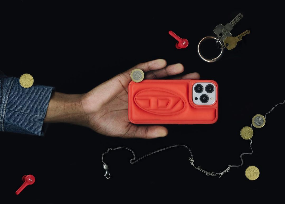 Hand holding a coral-colored Diesel brand cell phone case with earbuds, keys, and coins against a dark background in a studio shot.