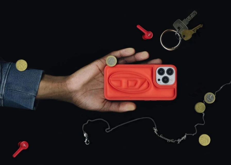 Hand holding a coral-colored Diesel brand cell phone case with earbuds, keys, and coins against a dark background in a studio shot.