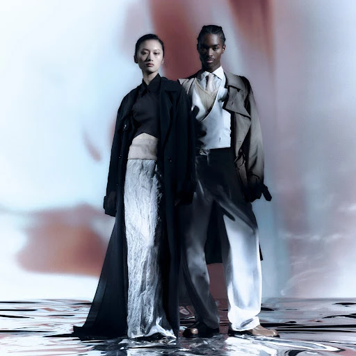 A stylish couple wearing coats stands on a reflective surface in a fashion-forward editorial setting with a blurred backdrop.