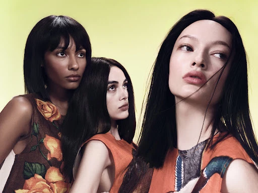 Three diverse fashion models pose in a studio with a light yellow backdrop, wearing patterned tops in a close-up shot. Editorial fashion.