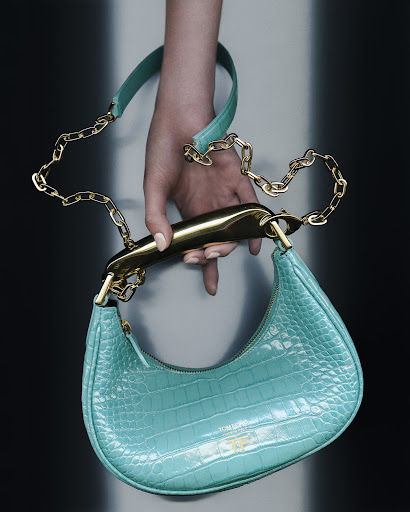 Close-up of a turquoise handbag with a gold handle and chain, held by a fair-skinned hand with manicured nails, against a neutral background.