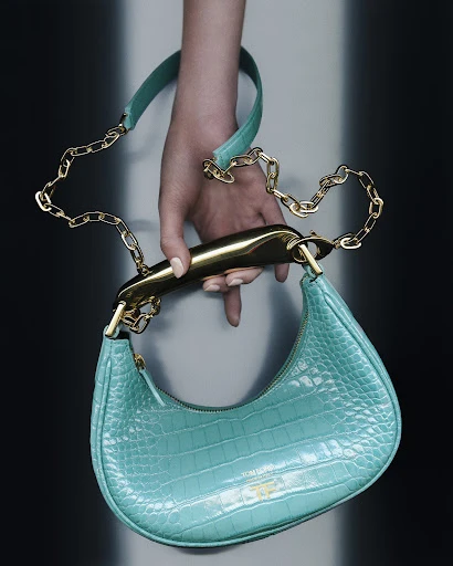 Close-up of a turquoise handbag with a gold handle and chain, held by a fair-skinned hand with manicured nails, against a neutral background.