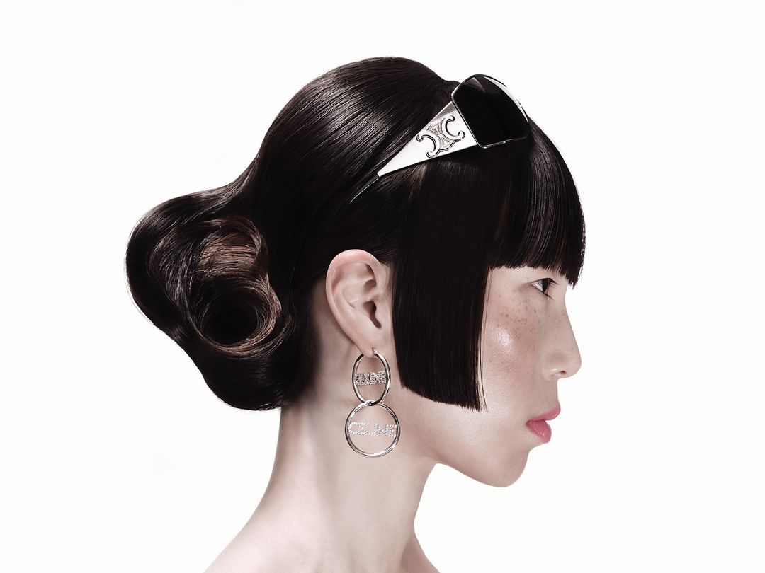 Profile portrait of a woman wearing statement earrings and designer sunglasses resting on dark pinned-up hair against white seamless backdrop.