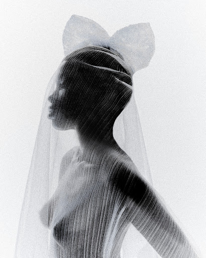 Fashion shot of a Black female model veiled in sheer fabric with a large bow, set against a bright white background in a studio.
