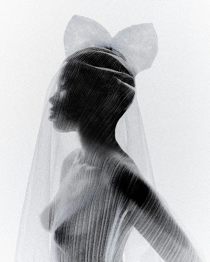 Fashion shot of a Black female model veiled in sheer fabric with a large bow, set against a bright white background in a studio.