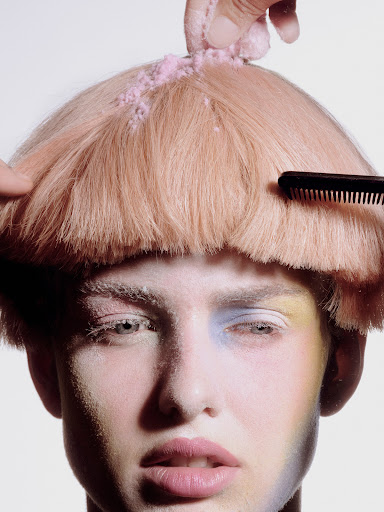 Close-up shot of a model getting styled with pink powder and comb, featuring pastel makeup and a fair complexion against a light backdrop.