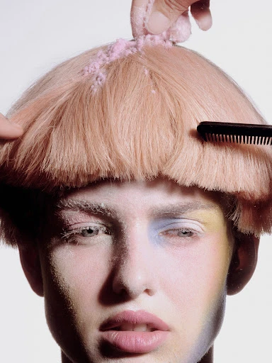 Close-up shot of a model getting styled with pink powder and comb, featuring pastel makeup and a fair complexion against a light backdrop.