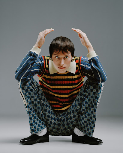 Adult male crouches indoors wearing a horizontally striped sweater, collared shirt, patterned pants, and black patent-leather shoes in a studio setting.