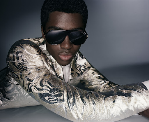 A stylish dark-skinned man in a gold-patterned jacket and sunglasses reclines in a studio with diffused lighting, showcasing a blend of fashion and portraiture.