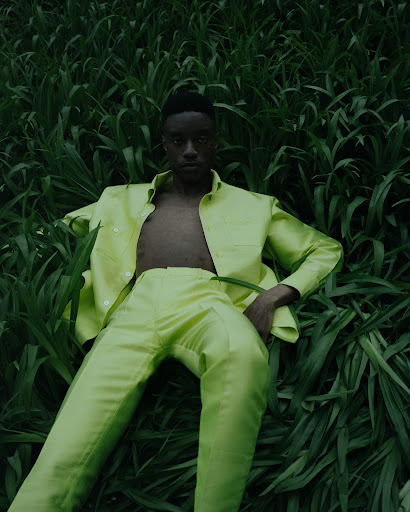Person in a light green suit reclining in a field of tall grass. Fashion editorial shot with natural lighting and a moody aesthetic.