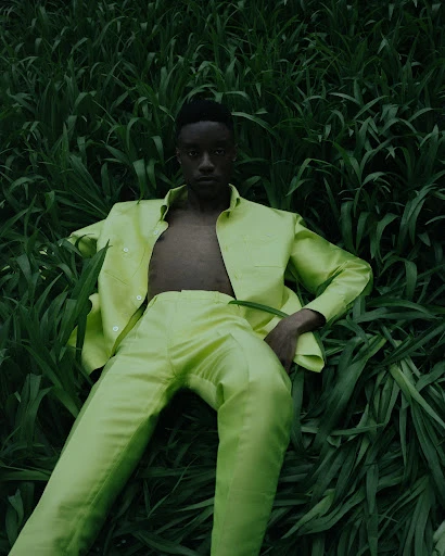 Person in a light green suit reclining in a field of tall grass. Fashion editorial shot with natural lighting and a moody aesthetic.
