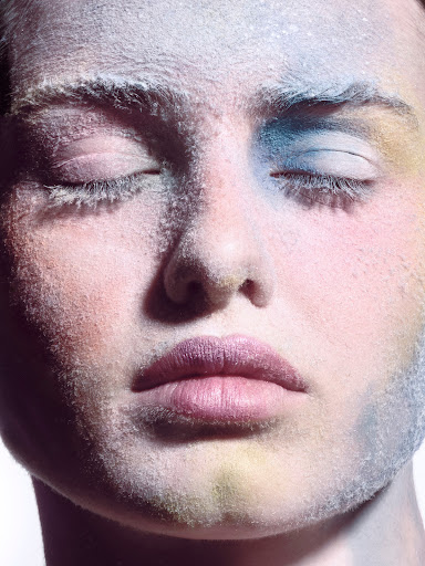 Close-up studio portrait of a fair-skinned adult female with eyes closed, face covered in multicolored powder paint.